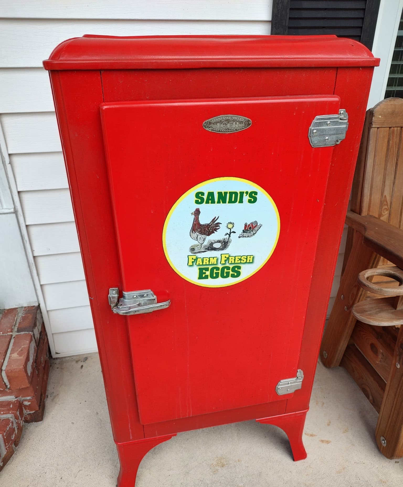 A restored red icebox painted with Sandi's Farm Fresh Eggs, a hen, a sunflower and a red barn