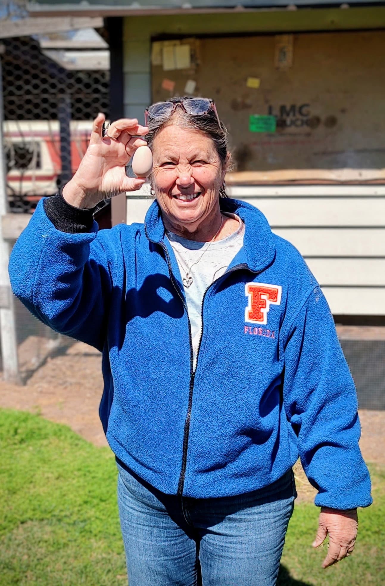 Sandi smiling and holding up a freshly collected egg outside the coop